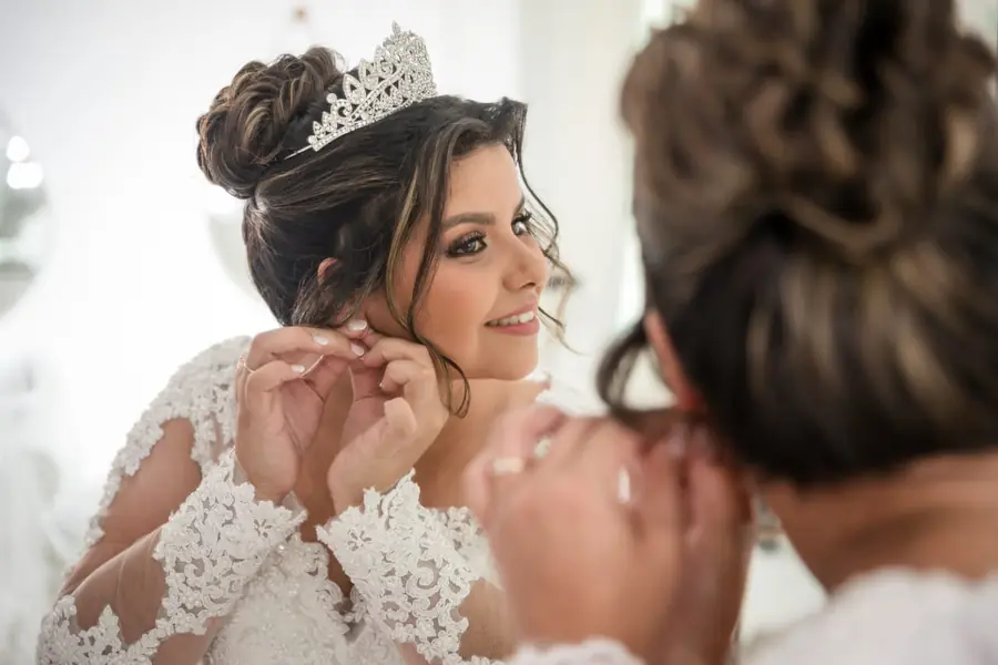 Noiva com tiara colocando os brincos em frente ao espelho no Dia da Noiva